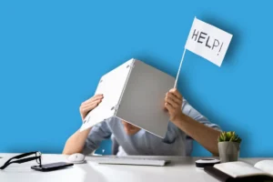 un travailleur caché sous son ordinateur de travail avec un drapeau HELP (aidez-moi, en anglais) à cause de sa surcharge de travail